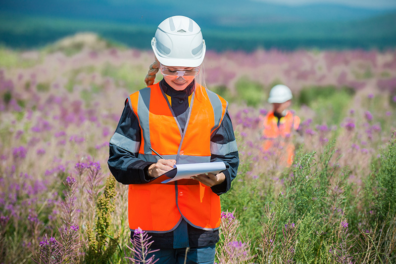 woman in white hard hat and prange safety vest carrying clipboard walking through field