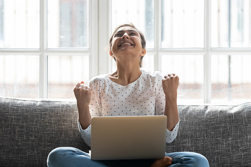 Overjoyed young Indian woman celebrating while sitting with computer on couch at home.